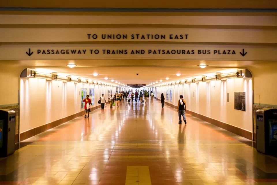 View looking down indoor concourse at Los Angeles Union Station with “To Union Station East” and “Passageway to Trains and Patsaouras Bus Plaza” signs.